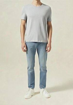 Homme portant un t-shirt gris clair à manches courtes, un jean bleu clair et des baskets blanches, debout devant un fond beige uni.