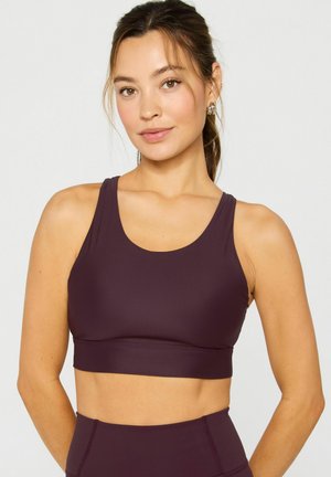 Frau mit zurückgebundenen Haaren, die einen ärmellosen, dunkelvioletten Sport-BH und passende, hoch taillierte Leggings trägt, steht vor einem schlichten Hintergrund.