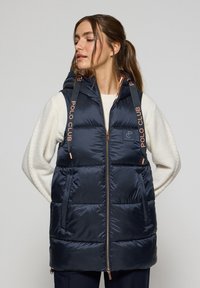 Veste sans manches matelassée bleu marine avec capuche, présentant des accents rose doré, une fermeture éclair à l'avant et deux poches latérales. Logo sur la poitrine gauche.