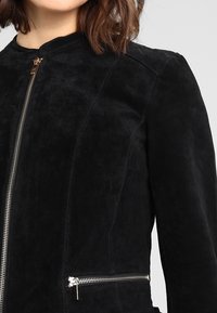 Žena v černé semišové bundě s předními a bočními stříbrnými zipy, viditelným švem na rameni a hnědými vlasy po ramena.