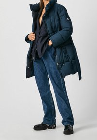 Marineblå pufferjacka med hög krage, fickor med dragkedja och kviltad struktur, kombinerad med mörkblå vida jeans och svarta klumpiga skor.