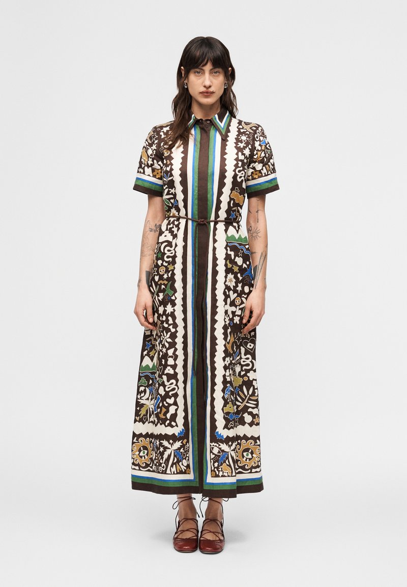 Γυναίκα που φοράει μακρύ, πουκαμισ οφόρεμα με μοτίβα σε καφέ, λευκό, πράσινο και μπλε χρώμα, συνδυασμένο με καφέ δερμάτινα παπούτσια με κορδόνια, σε απλό φόντο.