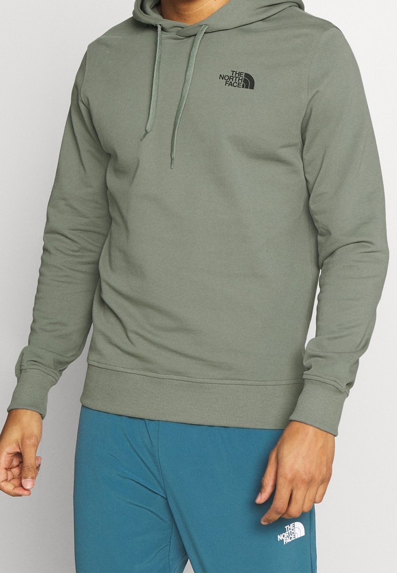 Homme portant un sweat à capuche vert olive et un pantalon bleu, tous deux avec le logo The North Face.