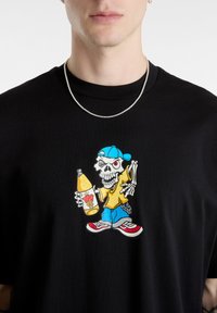 Black cotton t-shirt featuring a colorful graphic of a skeleton character in a yellow shirt, blue pants, and a blue cap, holding a drink.