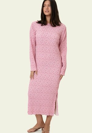 Frau trägt ein langes, rosa, geripptes Kleid mit kleinen roten Blumenmustern, langärmlig, mit einem seitlichen Schlitz, steht vor einem einfarbigen Hintergrund.