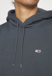 Primer plano de una persona que lleva una sudadera con capucha gris oscuro con cordones y un pequeño logo rectangular rojo, blanco y azul en el pecho.