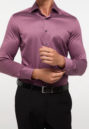 Uomo che sistema il polso di una camicia aderente viola a maniche lunghe infilata in pantaloni neri con cintura nera.