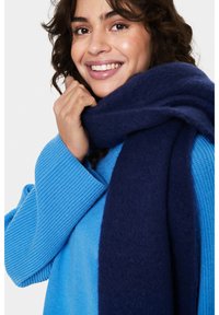 Pull bleu à côtes associé à une écharpe bleu foncé. L'écharpe a une apparence douce et texturée, drapée doucement autour du cou.