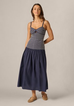 Kvinde står iført en ærmeløs navyblå kjole med hvide striber på toppen og ensfarvet nederdel, brune flade sko og øreringe med hul.