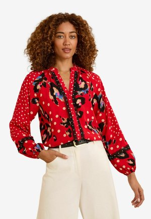 Blusa rossa con pois e un motivo floreale, con bottoni sul davanti, dettagli neri e maniche a palloncino. Indossata con pantaloni color crema.
