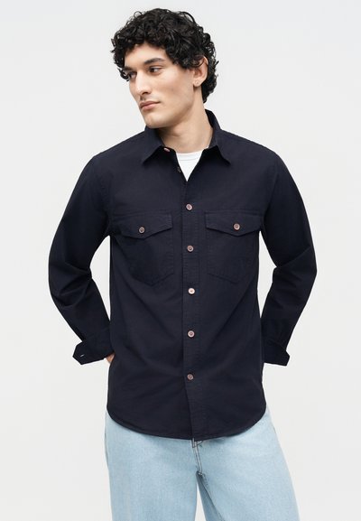 Νέος άντρας με σγουρά μαλλιά, φορώντας ένα σκούρο πουκάμισο με τσέπες στο στήθος και ανοιχτόχρωμο τζιν, στέκεται με τα χέρια πίσω από την πλάτη.