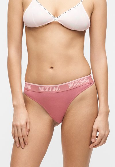 Γυναίκα φοράει λευκό σουτιέν με λεπτές λουρίδες με λογότυπο και ροζ εσώρουχα μπικίνι Moschino με λογότυπο, εμφανιζόμενη από τη μέση του κορμού μέχρι το επάνω μέρος των μηρών.