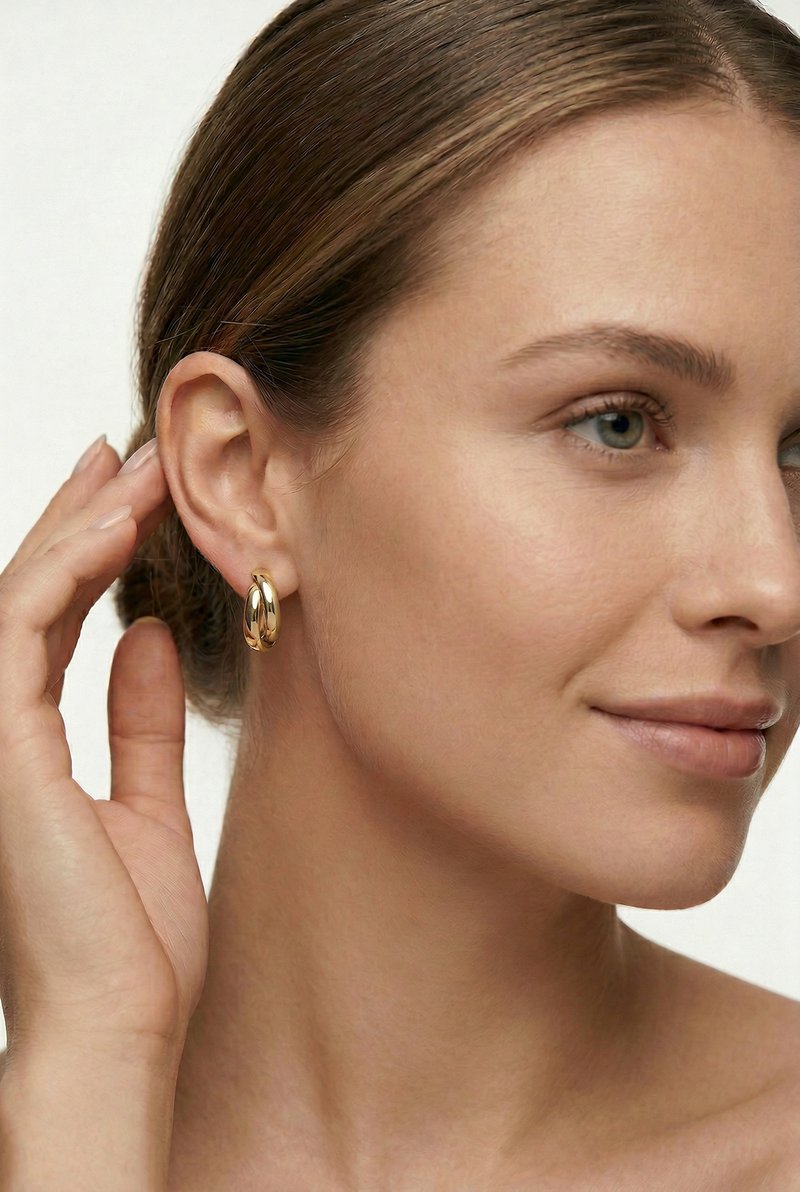 Mujer de piel clara y ojos azules tocándose la oreja, con un pequeño aro grueso de oro contra un fondo neutro.
