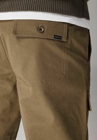 Bruine cargobroek van een textuurstof met een achterzak en een klein blauw label. Naai-details zijn zichtbaar aan de omtrek van de zak.