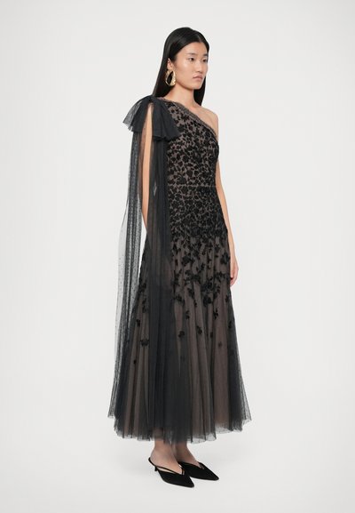 Femeie îmbrăcată într-o rochie din tul negru cu un umăr, cu broderie florală, o fundă pe umăr, cercei aurii în formă de cerc și pantofi negri cu vârful ascuțit.