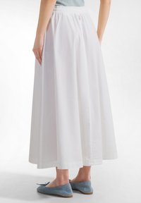 Kvinde iført en hvid lang nederdel og lyseblå flade sko, vist bagfra mod en ensartet hvid baggrund.