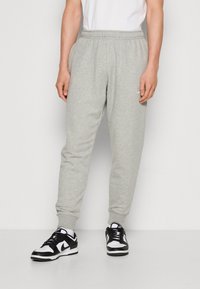 Hombre vestido con pantalones de chándal gris claro, calcetines blancos y zapatillas Nike en blanco y negro, de pie contra un fondo blanco simple.