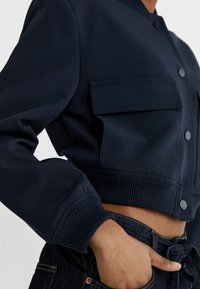Navy cropped bomber jacket with two front pockets, ribbed cuffs, and a button closure, featuring a smooth fabric texture.