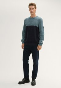 Chunky gestrickter Pullover in blauem Verlauf mit gerippten Bündchen und Saum. Kombiniert mit dunklen Hosen und schwarzen Schnürstiefeln, steht vor einem neutralen Hintergrund.