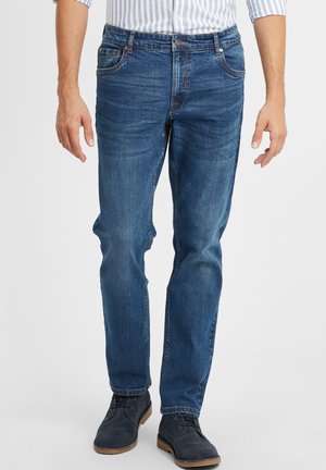 Uomo che indossa jeans slim-fit blu, stivali di camoscio blu navy e una camicia bianca con righe blu, in piedi su uno sfondo chiaro.