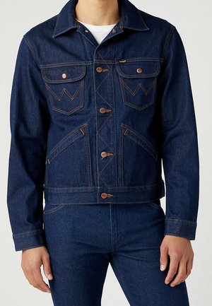 Personne portant une veste en denim bleu foncé avec fermeture à boutons et un pantalon en denim assorti sur un fond clair uni.