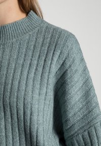 Primo piano di una persona che indossa un morbido maglione grigio-blu chiaro a coste con un collo alto e rotondo su uno sfondo semplice.
