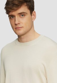Pull beige clair avec un col rond, un col côtelé et des manches longues. Texture lisse, design minimal et coupe ajustée.