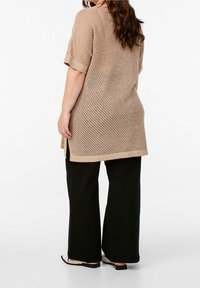 Beige gebreide tuniek met korte mouwen, featuring een losse, open weefstructuur en zijsplitten. Gecombineerd met wijde zwarte broeken en platte sandalen.