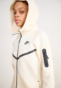 Suenadeira com fecho éclair na cor creme, apresentando um fecho e uma faixa preta no peito, com um logo preto da Nike e um bolso com fecho na manga.