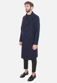 Cappotto in lana blu navy doppiopetto con colletto a scendenti, tasche frontali e bottoni. Modello che indossa pantaloni neri slim-fit e scarpe nere.