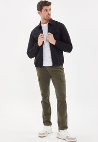 Zwarte ritsjack met elastische manchetten, olijfgroene cargobroek met zijzakken en witte sneakers met grijze accenten. Model staat voor een neutrale achtergrond.