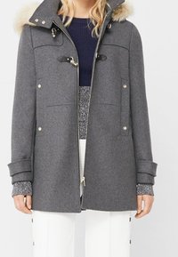Parka grise avec capuche en fourrure, présentant des accents dorés, fermeture à glissière sur le devant et poches latérales. Matériau texturé avec des manches à revers.
