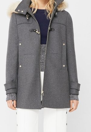 Parka grise avec capuche en fourrure, présentant des accents dorés, fermeture à glissière sur le devant et poches latérales. Matériau texturé avec des manches à revers.