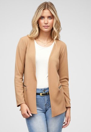 Hellbraune, strukturierte Jacke mit entspannter Passform, ausgestattet mit zwei Reißverschlusstaschen, über einem weißen Oberteil getragen und kombiniert mit hellblauen Jeans.