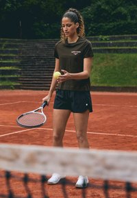 Camicia marrone a maniche corte con testo bianco, pantaloni corti neri, che tiene una pallina da tennis gialla e una racchetta, in piedi su un campo da tennis in terra battuta.