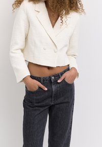 Weißes, strukturiertes, doppelt knöpfiges, kurzes Blazer mit Reverskragen, kombiniert mit hoch taillierten, dunklen Denim-Jeans. Hände in den Taschen.