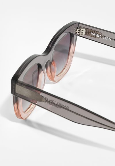 Lunettes de soleil avec des montures surdimensionnées, transparentes grises et roses. Verres dégradés et logo de la marque gravé sur la branche. Design élégant et moderne.
