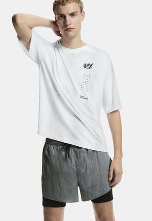 Bărbat tânăr purtând un tricou alb "RUN Swiss Engineering" și pantaloni scurți de alergare gri în straturi, pozând cu o mână în spatele capului.