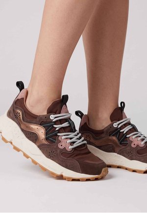 Coppia di scarpe sportive marroni e rosa con suole bianche e lacci grigi indossate a piedi nudi su uno sfondo semplice.