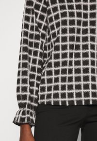 Schwarz-weiß gemusterte Bluse mit einem Karodesign. Verfügt über puffige Ärmel mit Rüschebündchen. Der Stoff hat eine weiche Textur.