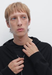 Schwarze Zip-Hoodie mit gerippten Bündchen, über einem schwarzen Shirt getragen. Das Model justiert den Reißverschluss vor einem neutralen Hintergrund. Kurzes blondes Haar und helle Haut.