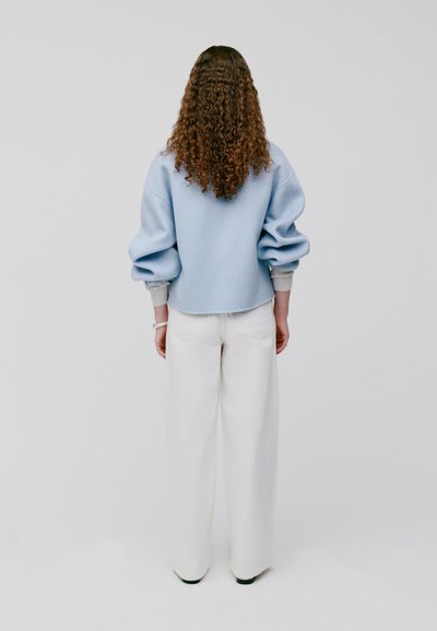 Lichtblauwe, oversized sweatshirt met opstaande mouwen en geribbelde manchetten, gecombineerd met witte wijde broekspijpen, gezien van achteren.