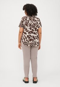 Model seen from behind, wearing a brown and cream spotted top, light beige pants, and green shoes against a plain white background.
