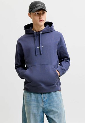 Jeune homme portant un sweat à capuche bleu marine, un jean bleu clair et une casquette noire, les mains dans les poches du sweat, regardant légèrement sur le côté.