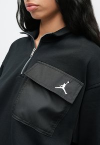 Κοντινό πλάνο ατόμου που φορά μαύρο φούτερ με φερμουάρ και μεγάλη τσέπη στο στήθος που έχει λογότυπο λευκού παίκτη μπάσκετ σε άλμα.