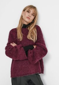 Vinröd mjuk tröja med vida ärmar och avslappnad passform, lager-på-lager över en svart polotröja, kombinerad med mörka byxor.