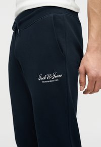 Δεξιά πλευρά ατόμου που φοράει μαύρο φόρμα παντελόνι με το κείμενο "Jack & Jones Archive Selection" κοντά στην τσέπη, λευκό πουκάμισο μερικώς ορατό.