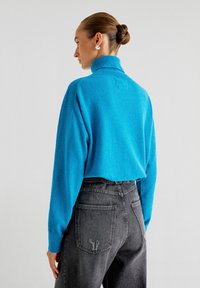Übergroßer Rollkragenpullover aus leuchtend blauem Strick, kombiniert mit hoch sitzenden, dunkelgrauen Jeans mit beschädigten Details an den Hintertaschen.