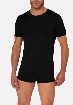 Mann trägt ein eng anliegendes schwarzes kurzärmeliges T-Shirt und schwarze Boxershorts vor einem schlichten hellen Hintergrund.