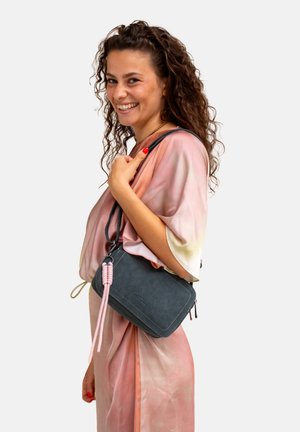 Blue suede crossbody bag with a pink braided accent, rectangular shape, and contrast stitching. The model wears a soft, gradient fabric.